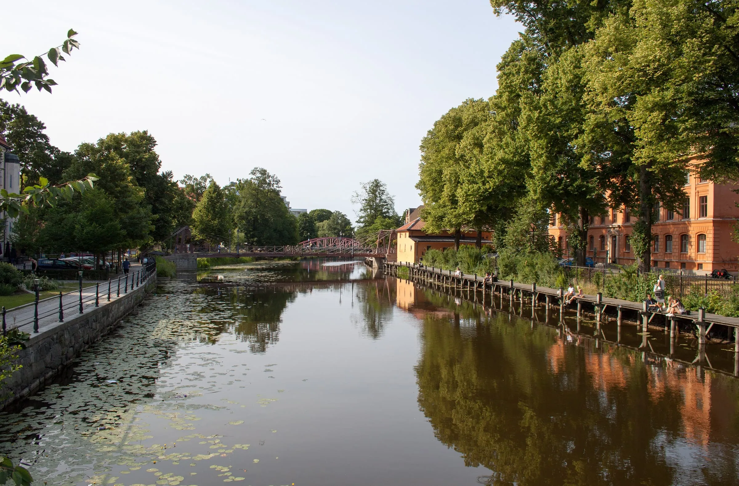 Fyrisån i centrala Uppsala en sommardag med lummiga träd, en röd bro i bakgrunden och människor som sitter på kajen.