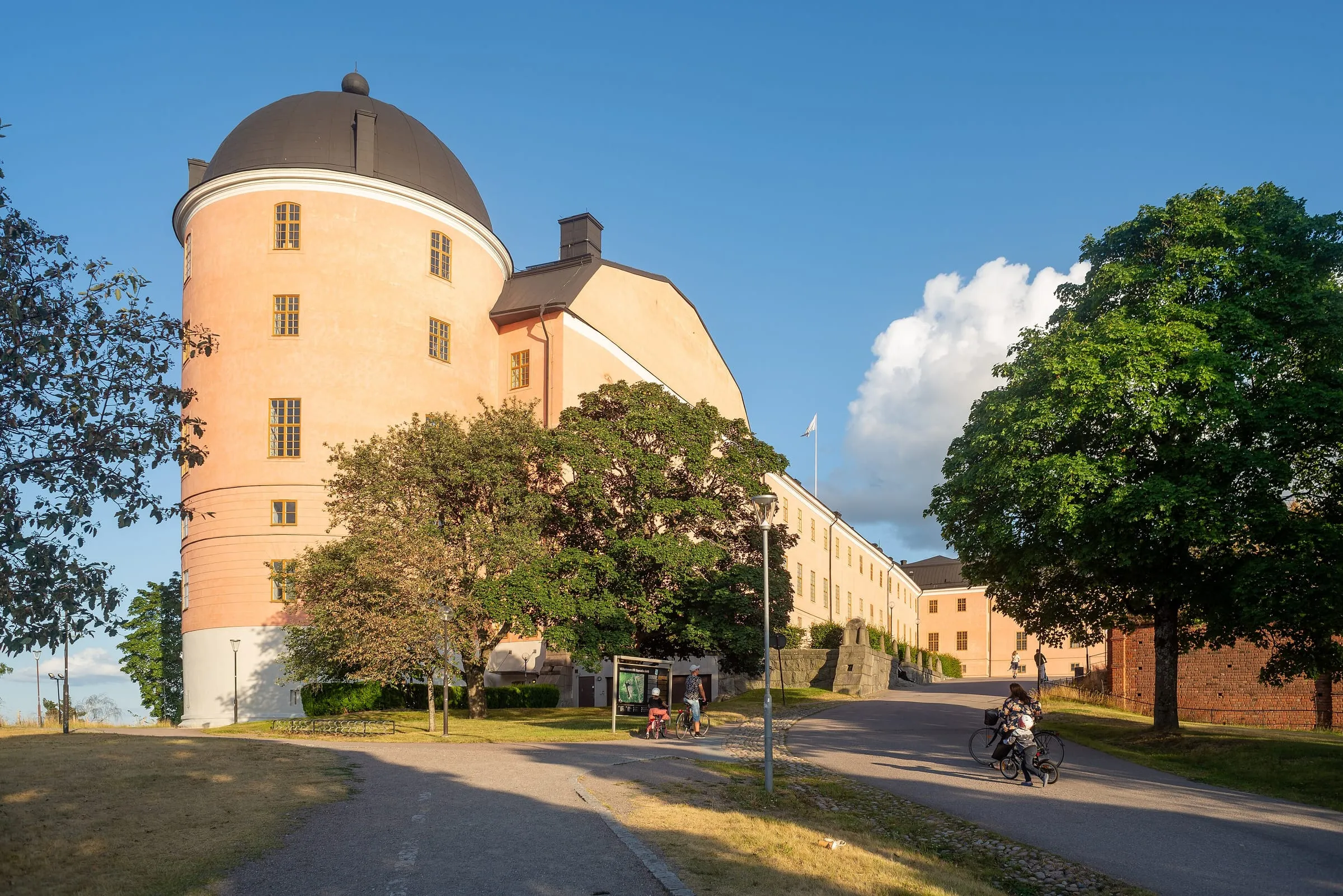 Uppsala slott en sommarkväll med rosa fasad, lummiga träd och en cyklist på vägen framför.
